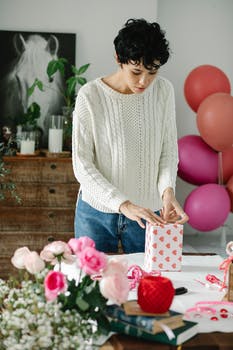 Focused young ethnic female millennial with curly dark hair in stylish outfit packing gift with wrapping paper while standing in room decorated with balloons and flowers
